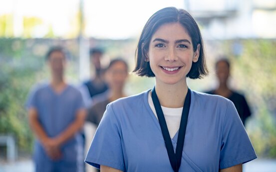 Junge Frau mit kurzen braunen Haaren im blauen medizinischen Kittel lächelt, unscharfe Gruppe in Berufskleidung im Hintergrund