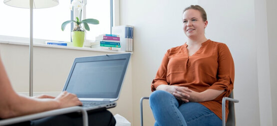 Zwei Personen im Gespräch, eine arbeitet am Laptop, die andere sitzt lächelnd im Stuhl