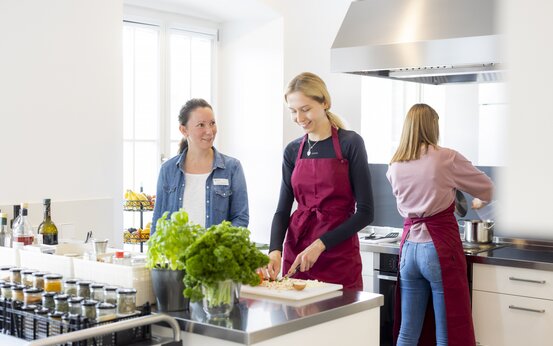 Drei Frauen kochen gemeinsam in moderner Lehrküche, schneiden Gemüse und rühren am Herd