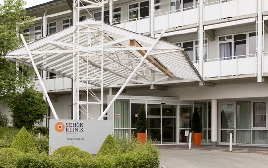 Haupteingang der Schön Klinik Roseneck mit Glasdach, großem Eingangsschild, automatischer Tür, gepflegtem Grün und Balkonen im Hintergrund