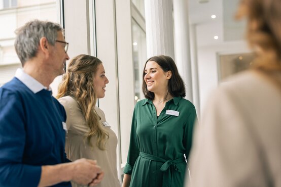 Zwei Frauen und ein Mann mit Namensschildern unterhalten sich lächelnd im modernen Klinikraum vor großer Fensterfront