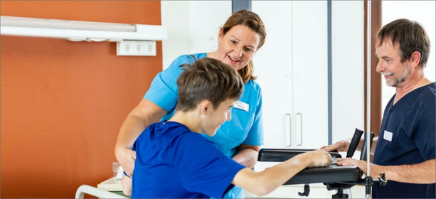 Zwei Fachkräfte im hellen Klinikflur unterstützen jungen Patienten in blauer Kleidung beim Gehen