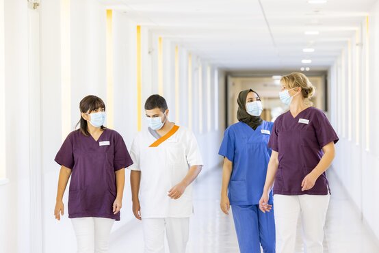 Vier Pflegekräfte im farbigen Kasack und Maske gehen gemeinsam durch hellen, modernen Klinikflur, lichtdurchflutete Fensterfront
