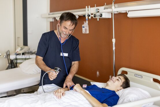 Pfleger im dunkelblauen Kasack misst Blutdruck bei jugendlichem Patienten im Klinikbett, beide freundlich, medizinische Geräte im Hintergrund