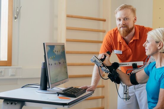 Physiotherapeut unterstützt Patientin bei Armtraining an Therapiegerät mit Bildschirm im modernen, hellen Klinikraum für Physiotherapie