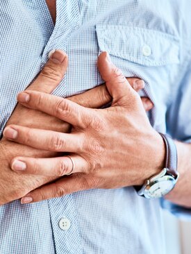 Mann in hellblauem Hemd hält sich mit beiden Händen die Brust, Symbolbild für Herzinfarkt oder akute Herzbeschwerden