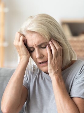 Ältere Frau sitzt auf dem Sofa und hält sich die Schläfen, zeigt Anzeichen von Kopfschmerzen