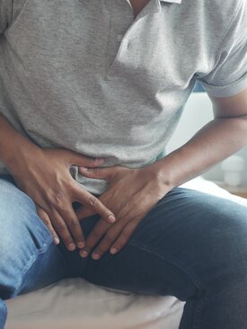 Mann sitzt auf dem Bett und hält sich mit beiden Händen den schmerzenden Unterbauch