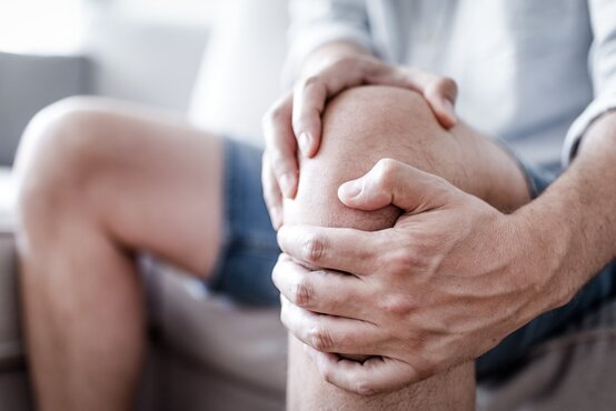 Mann hält sich schmerzhaft das rechte Knie, sitzt auf Sofa in Freizeitkleidung