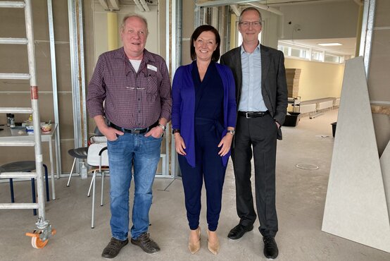 Drei Personen stehen in unfertigem Innenraum mit Metallständern und Baumaterial, Frau im Hosenanzug, zwei Männer in Hemd und Anzug