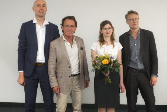 Junge Frau mit Blumenstrauß steht lächelnd mit drei Kollegen in Businesskleidung vor weißer Wand 