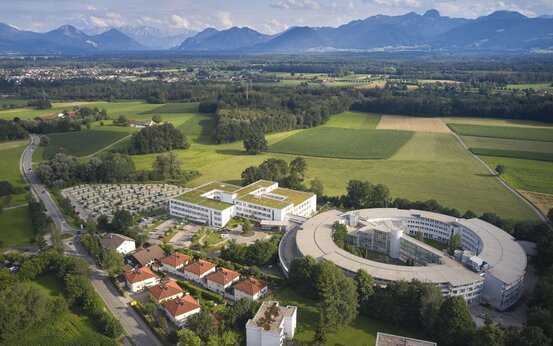 Luftaufnahme der Schön Klinik Bad Aibling Harthausen mit rechteckigem und rundem Bau, umgeben von Grünflächen und Bergen im Hintergrund