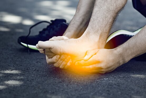 Person mit Sportschuhen sitzt auf Asphalt und hält sich schmerzerfüllt den linken Fuß mit Fokus auf Mittelfußbereich