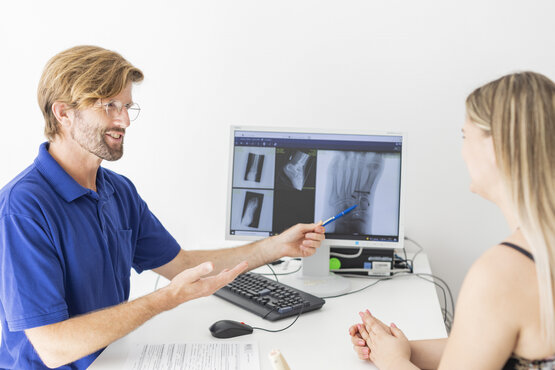 Orthopäde erklärt Patientin digitale Röntgenaufnahme des Fußes am Computerbildschirm in moderner Arztpraxis