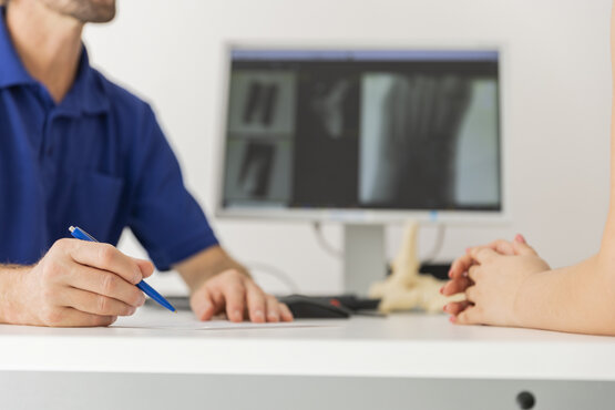 Zwei Personen am Schreibtisch in medizinischer Praxis, Monitor mit unscharfen Röntgenaufnahmen im Hintergrund