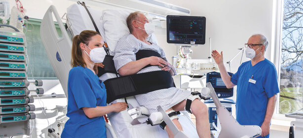 Älterer Patient mit Maske liegt fixiert im modernen Krankenhausbett, betreut von Krankenschwester und Arzt, Medizintechnik am Bett