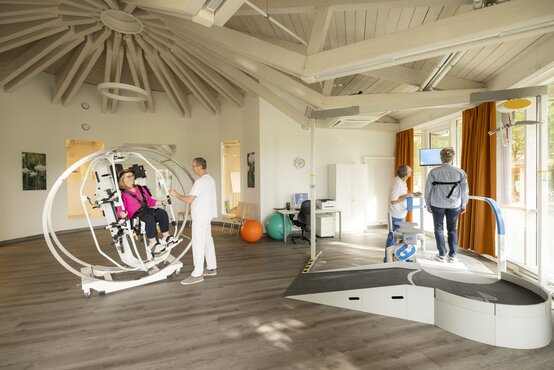 Therapieraum mit Patientin im Bewegungstrainer und Patient am Laufband, beide betreut von Therapeuten