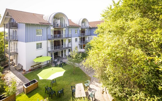 Moderner Gebäudekomplex mit drei Etagen, gepflegtem Garten, Sitzgruppen, Sonnenschirmen und viel Grün bei Sonnenschein