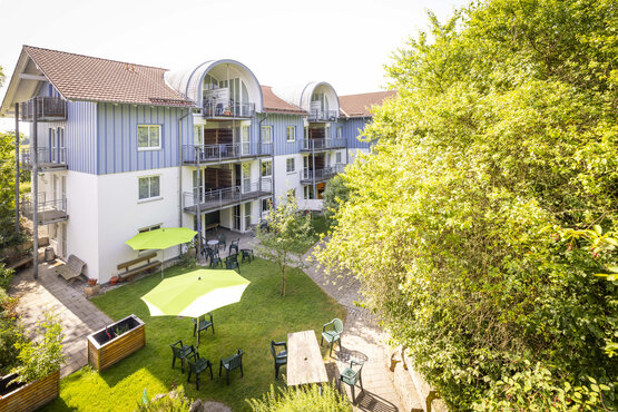 Moderner Gebäudekomplex mit drei Etagen, gepflegtem Garten, Sitzgruppen, Sonnenschirmen und viel Grün bei Sonnenschein