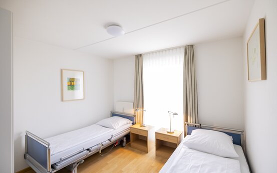 Helles, barrierefreies Pflegezimmer mit zwei höhenverstellbaren Betten, Nachttischen, großen Fenstern