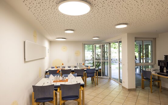 Heller Speisesaal im Alzheimer-Therapiezentrum mit gedeckten Holztischen, blauen Stühlen und großen Fenstern