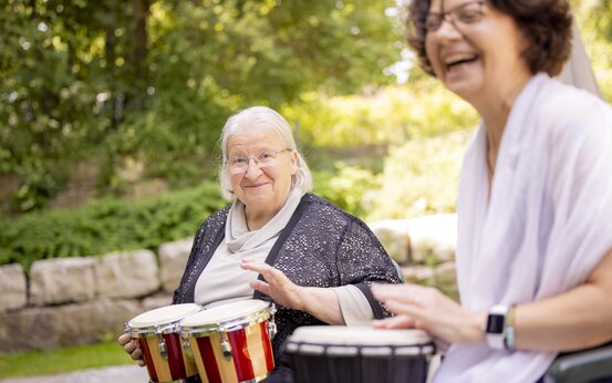 Zwei Frauen sitzen draußen vor Steinmauer und spielen Trommeln, eine hält Bongos, die andere spielt Djembe, beide lächeln