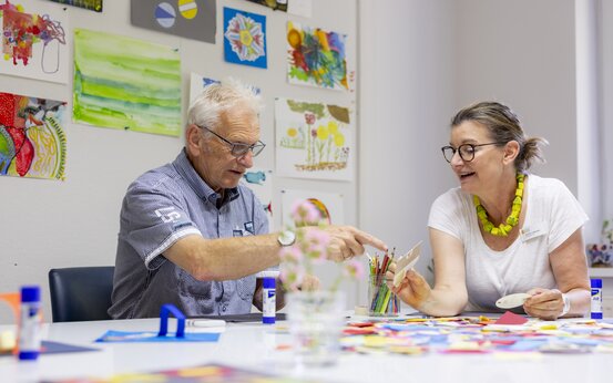 Zwei Personen sitzen im hellen Raum eines Alzheimer-Therapiezentrums an einem Tisch und machen gemeinsam Gestaltungstherapie