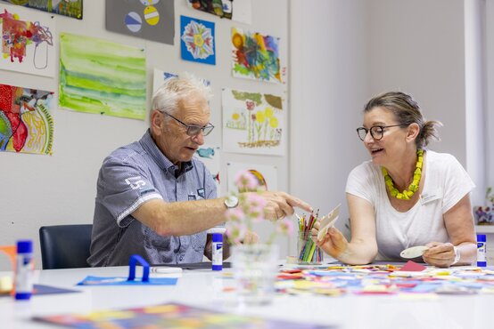 Zwei Personen sitzen im hellen Raum eines Alzheimer-Therapiezentrums an einem Tisch und machen gemeinsam Gestaltungstherapie