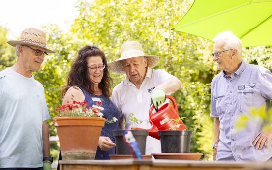 Vier ältere Erwachsene machen gemeinsam Gartenarbeit an Holztisch mit Blumentöpfen, eine Person gießt Pflanzen