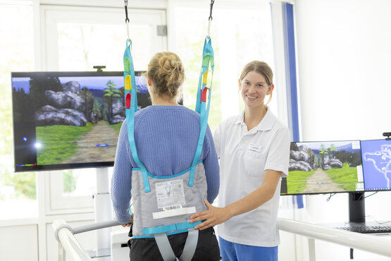 Physiotherapeutin unterstützt Patientin bei Gangrehabilitation mit Gehtrainer und Sicherheitsgurt, virtuelle Laufumgebung auf Bildschirmen