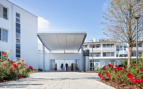 Moderne Klinik Außenansicht mit Haupteingang, weißes Gebäude, Glas-Eingangstür, Blumenbeete, blauer Himmel