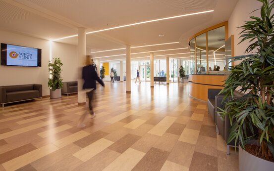 Helle Eingangshalle mit bodentiefen Fenstern, Empfangspult, Sitzbänken und großen grünen Pflanzen