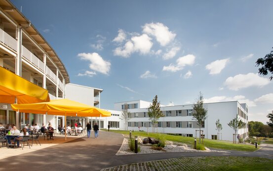 Barrierefreie Klinik mit weißer Fassade, gelbe Sonnenschirme, Menschen sitzen draußen an Tischen, grüne Rasenflächen, blauer Himmel