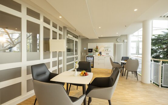 Helle, moderne Lounge mit Holzboden, großen Fenstern, grauen Sesseln und Küchenzeile mit Kaffeemaschine und Blumenstrauß