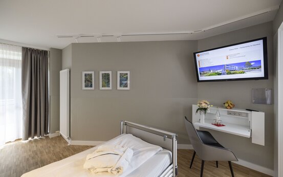 Modernes Klinik-Patientenzimmer mit großem Fenster, Schreibtisch, Flachbild-TV, Blumen und frischem Obst