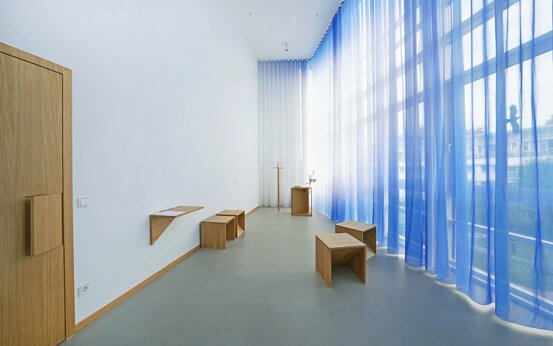 Heller, barrierefreier Andachtsraum mit Holzkreuz, bodentiefen Fenstern, weißen und blauen Vorhängen