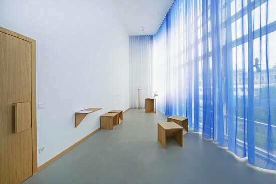 Heller, barrierefreier Andachtsraum mit Holzkreuz, bodentiefen Fenstern, weißen und blauen Vorhängen