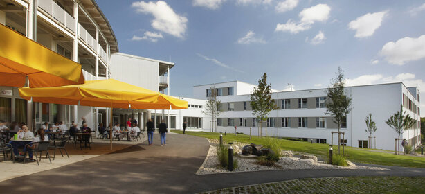 Moderner Klinikkomplex mit Terrasse, gelben Sonnenschirmen und Menschen im gepflegten Außenbereich