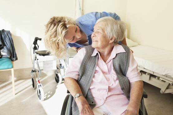 Junge Pflegekraft beugt sich freundlich zu älterer Frau im Rollstuhl, helles barrierefreies Pflegezimmer mit Pflegebett und Rollator