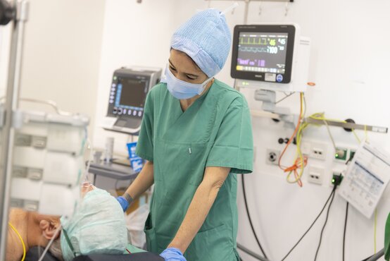 Medizinische Fachkraft in grüner OP-Kleidung und Mundschutz betreut Patient im Krankenhaus, medizinische Geräte im Hintergrund