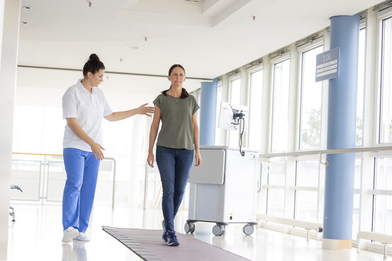Frau macht Geh-Test auf Gangteppich begleitet von Physiotherapeutin, medizinisches Analysegerät im Hintergrund