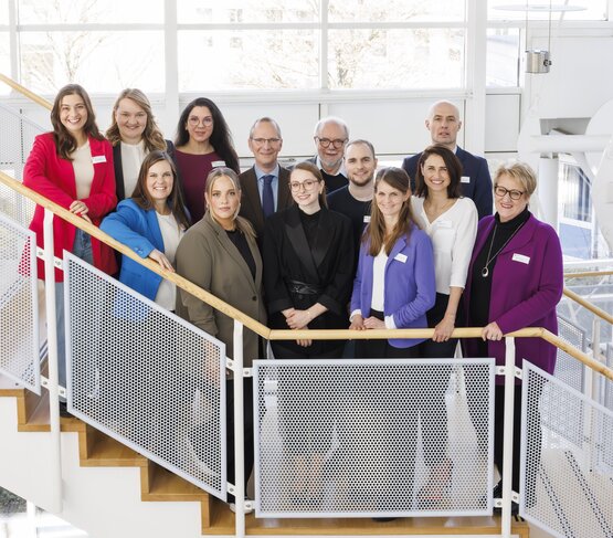 Dreizehnköpfiges Team in Business-Kleidung mit Namensschildern auf Treppe vor großen Fenstern