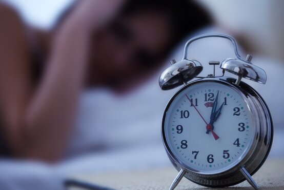 Silberner Wecker zeigt Uhrzeit auf Nachttisch, im Hintergrund liegt müde Person im Bett