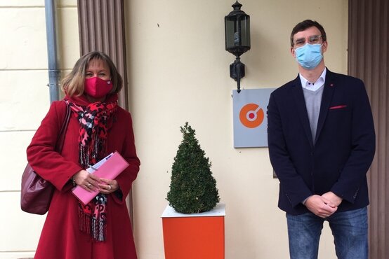 Frau mit rotem Mantel und Mann im blauen Sakko mit Masken vor Klinik-Eingang