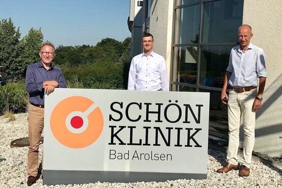 Drei Männer vor dem Eingang der Schön Klinik Bad Arolsen neben großem Firmenschild mit Kliniklogo