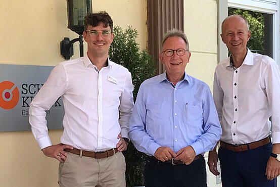 Drei lächelnde Männer in hellen Hemden vor dem Eingang der Schön Klinik neben dem Klinik-Schild