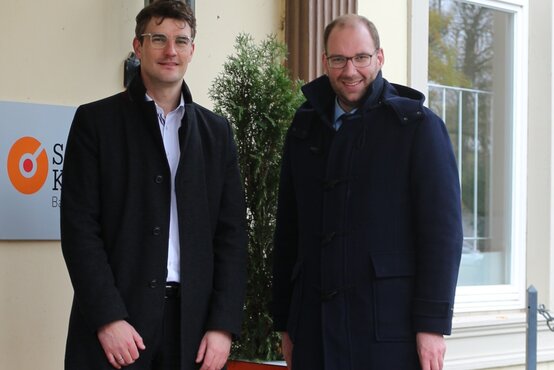 Zwei Männer mit Brille und dunklen Mänteln vor dem Eingang der Schön Klinik Bad Arolsen