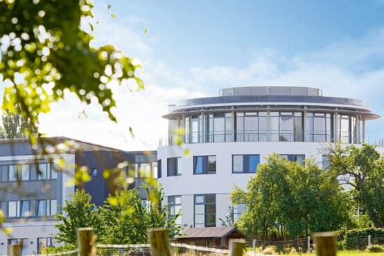 Modernes, rundes Klinikgebäude mit Glasfassade, umgeben von grünen Bäumen und Sträuchern