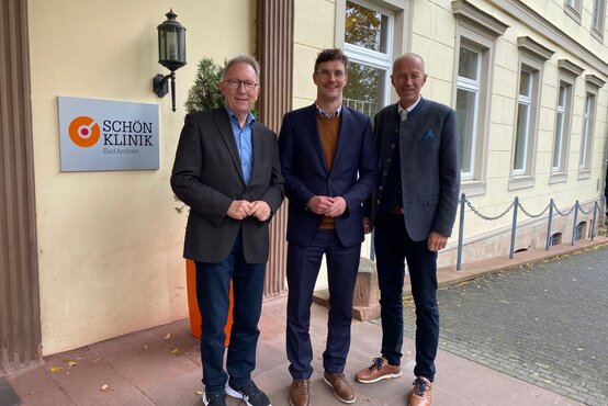 Drei Männer in Business-Kleidung vor dem Eingang der Schön Klinik Bad Arolsen neben Klinik-Schild an heller Hauswand