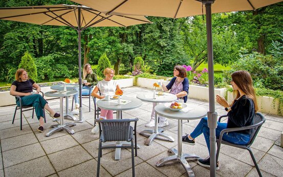 Fünf Frauen sitzen entspannt an runden Tischen auf sonniger Terrasse mit Kaffee, Kuchen und Blumen, umgeben von grüner Natur